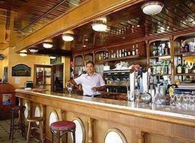 Zona de bar con un cantinero, mostrador de madera, taburetes y estantes llenos de botellas.