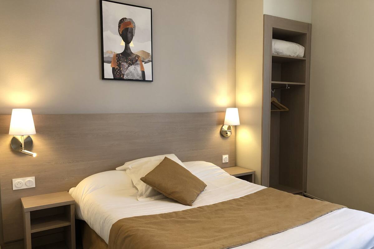 A hotel room with a bed, two bedside tables, a wall-mounted lamp, a coat closet, and a framed picture on the wall.