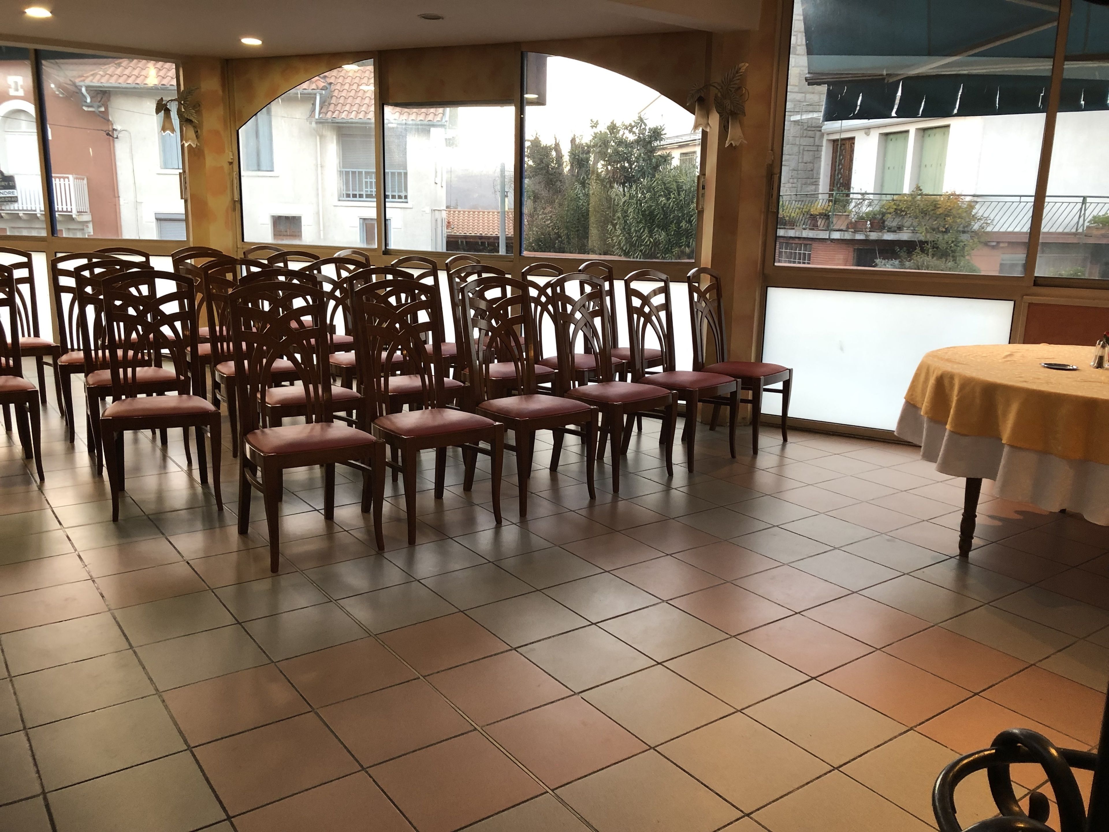 Una habitación con varias sillas de madera dispuestas en filas, ventanas amplias que dan a vistas de edificios y árboles, y una mesa con mantel amarillo.
