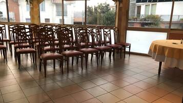 Una habitación con varias sillas de madera dispuestas en filas, ventanas amplias que dan a vistas de edificios y árboles, y una mesa con mantel amarillo.