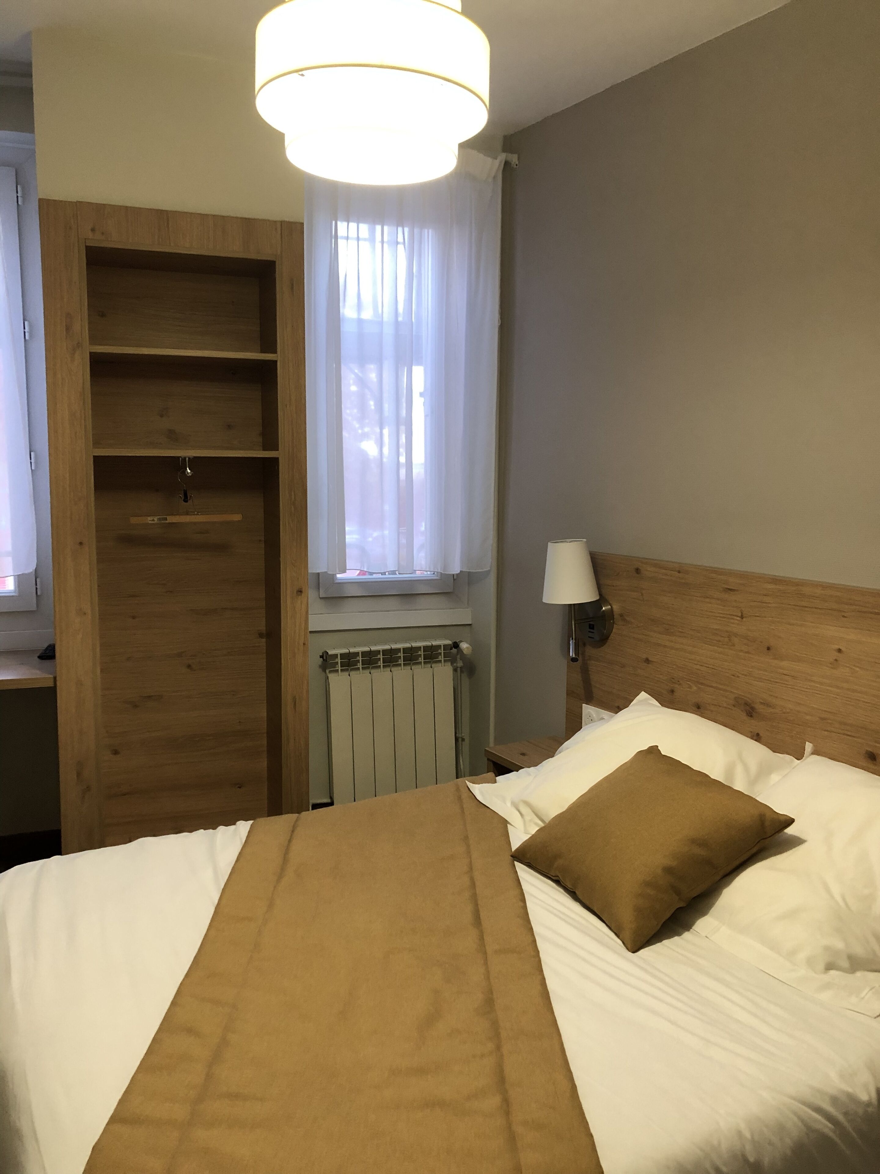 Una camera d'albergo con un letto, un armadio in legno, un termosifone, una finestra con tende e una lampada.