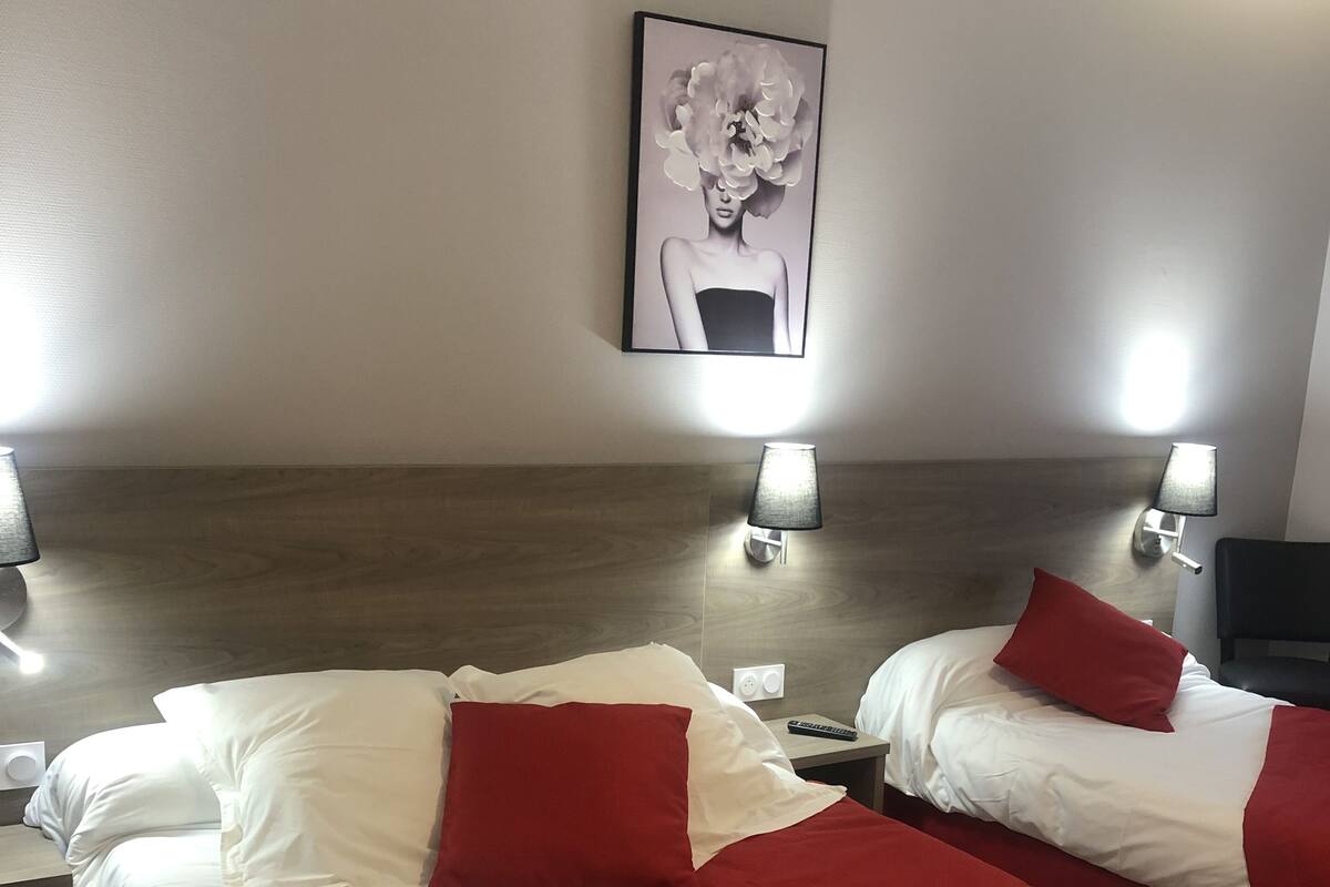 A hotel room with two beds, each with white and red bedding, a wooden headboard, a wall-mounted lamp, and a framed artwork above the beds.