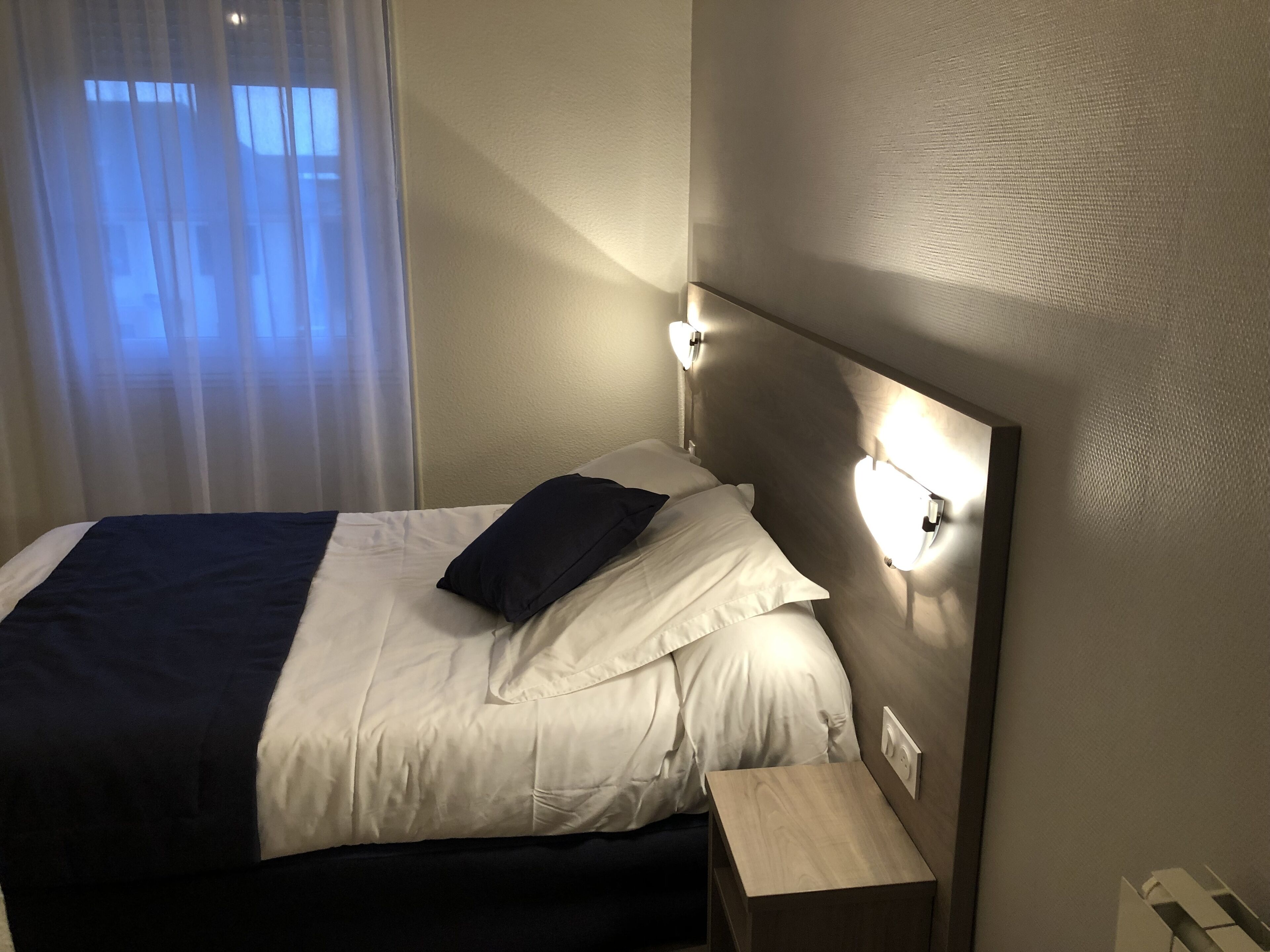 A neatly made bed with white linens and a dark blue blanket, a bedside table, and wall-mounted reading lights.