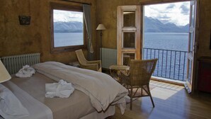 Een hotelkamer met een bed, een stoel, een tafel en uitzicht op een meer en bergen door een geopende deur.