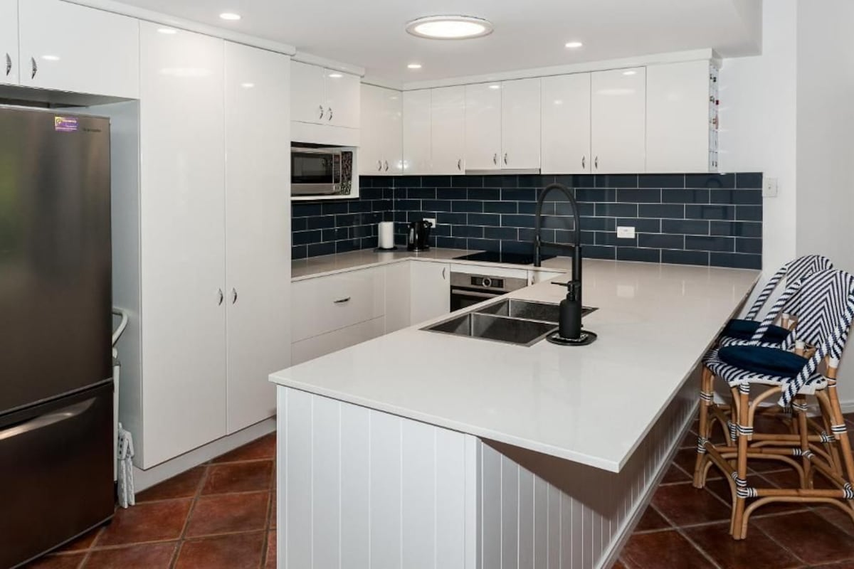 A modern kitchen with white cabinetry, a stainless steel refrigerator, a microwave, a sink, and a dining area with wicker chairs.