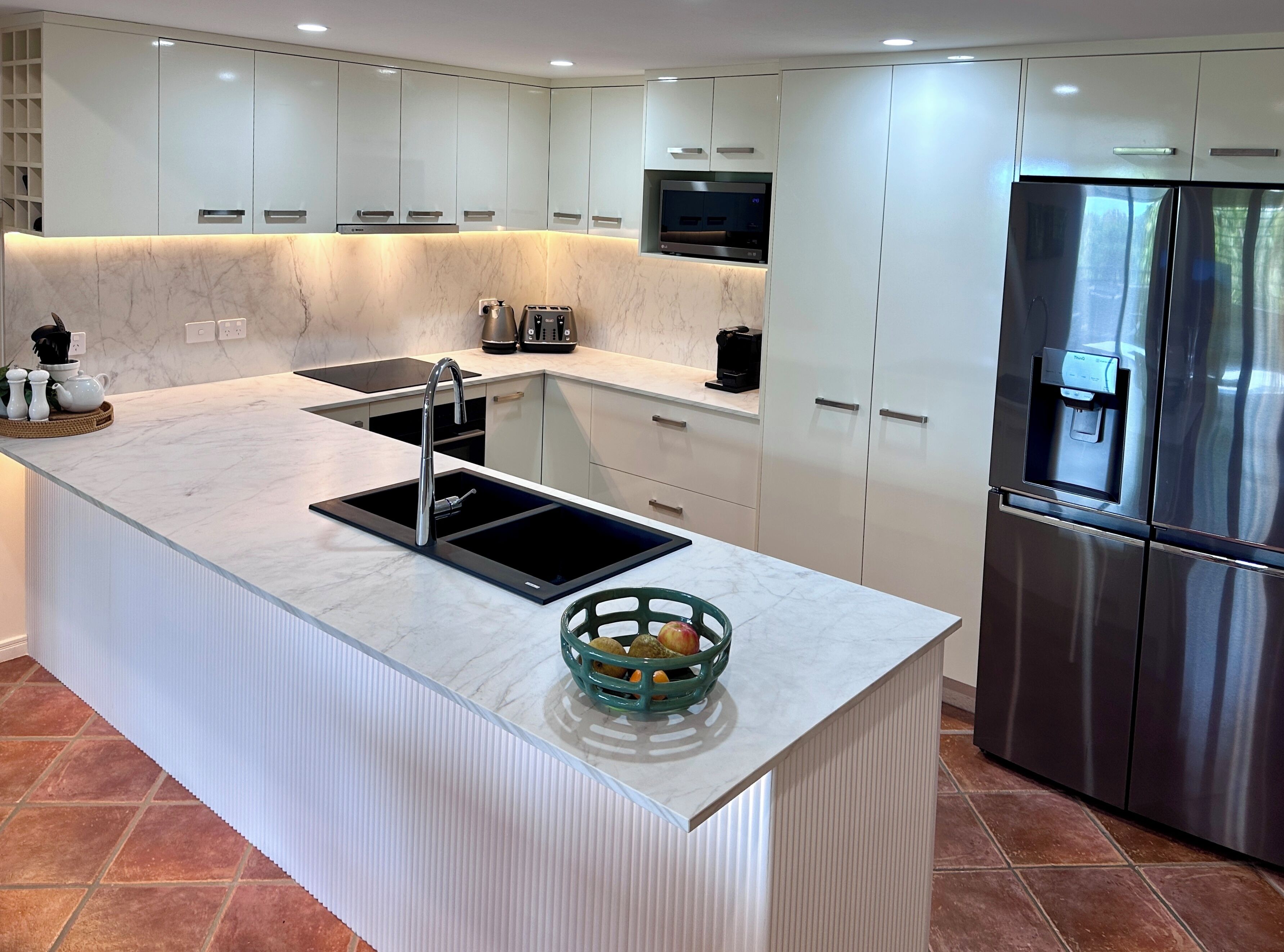 Une cuisine moderne avec des meubles blancs, un plan de travail en marbre, un évier encastré, un réfrigérateur en acier inoxydable et un petit panier de fruits posé sur l’îlot central.