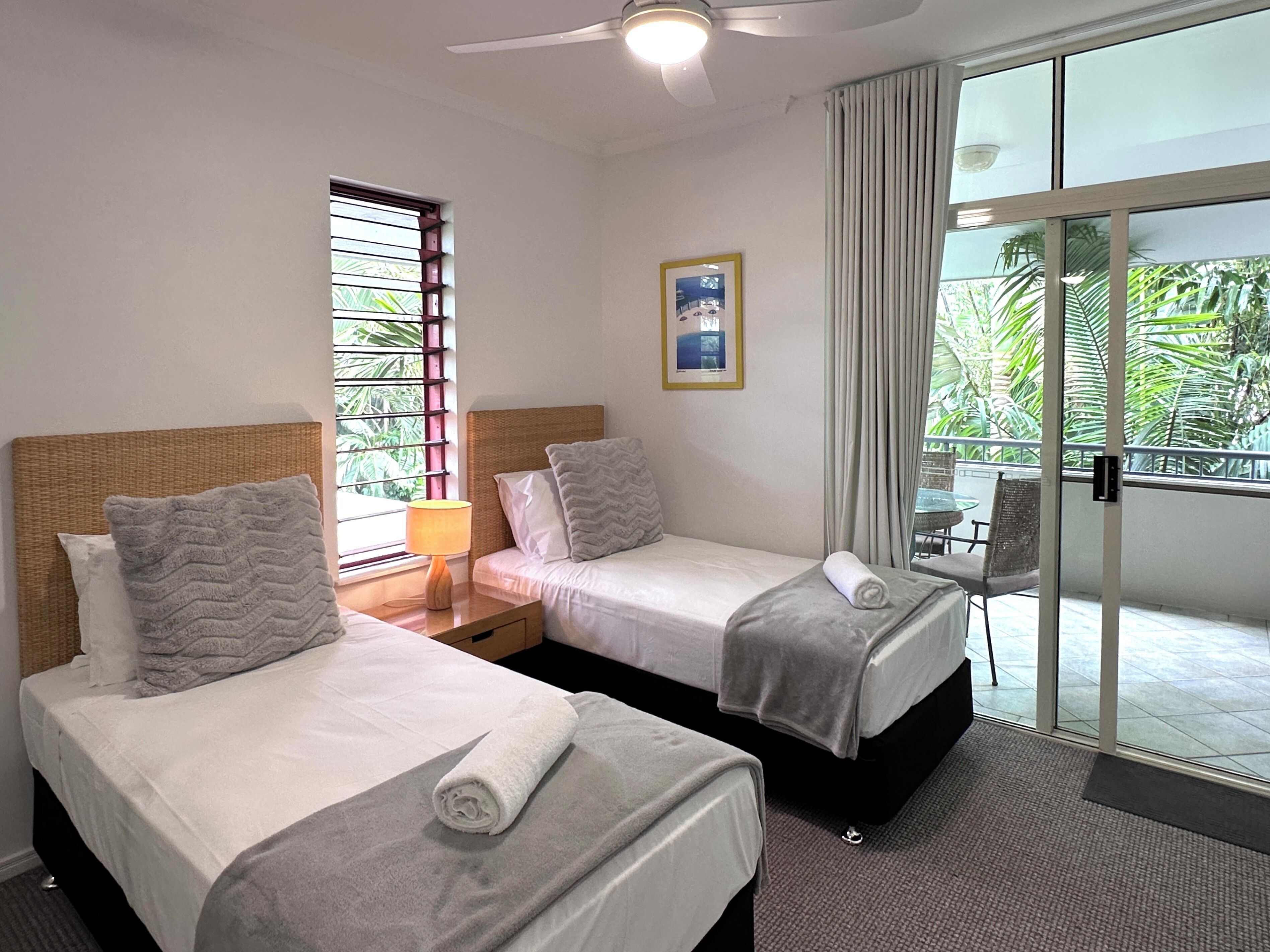 Une chambre d’hôtel avec deux lits, une fenêtre avec des stores, un ventilateur de plafond, un tableau au mur et un balcon donnant sur un paysage verdoyant.