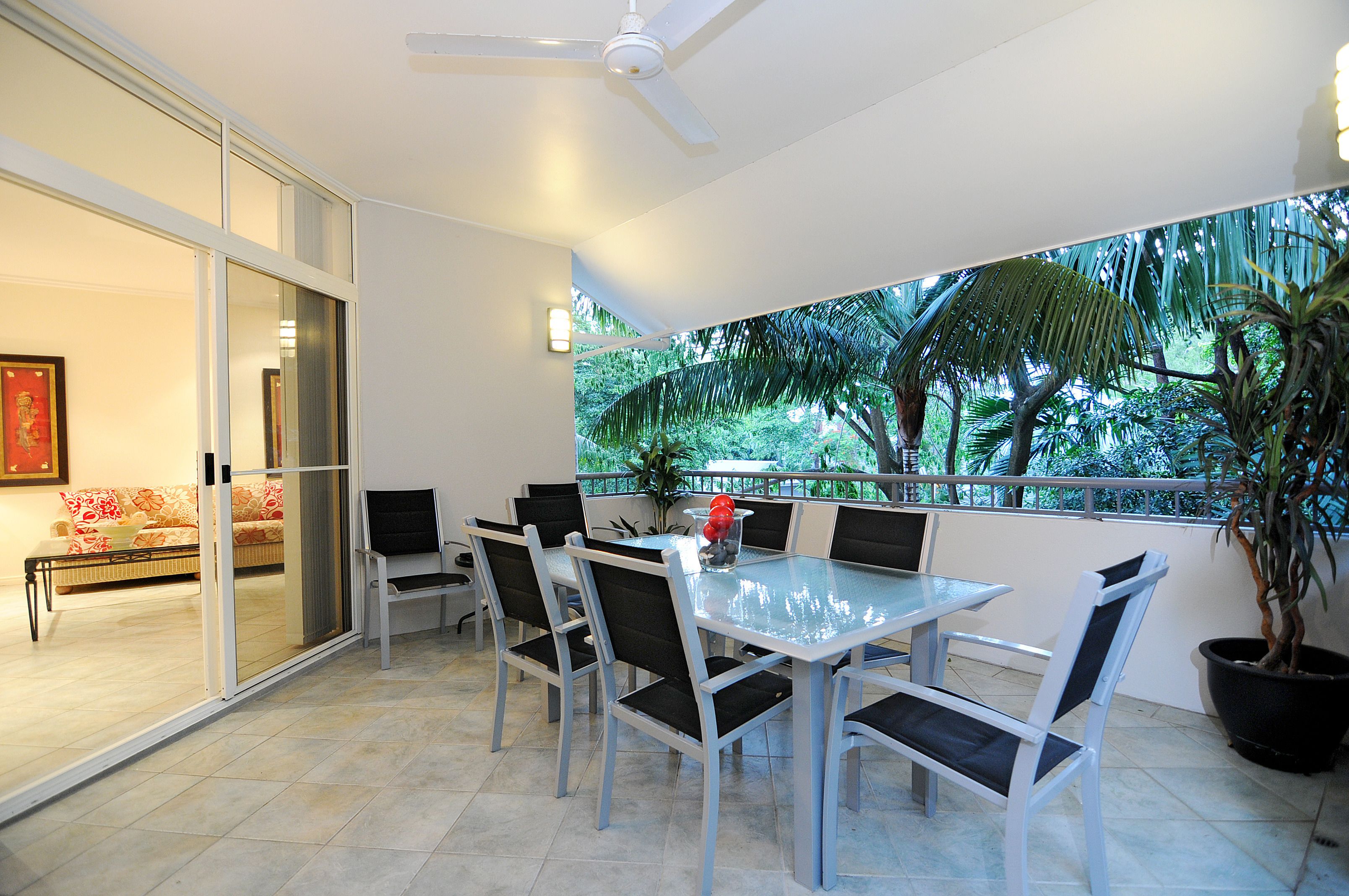 Zona pranzo con tavolo e sedie in vetro, ventilatore a soffitto e vista sul verde attraverso una porta di vetro.