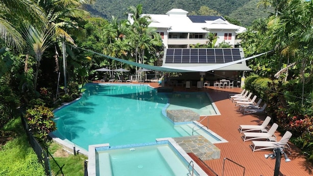 A pool area with a wooden deck, lounge chairs, and a building with solar panels in the background. - Oasis at Palm Cove (Palm Cove)
