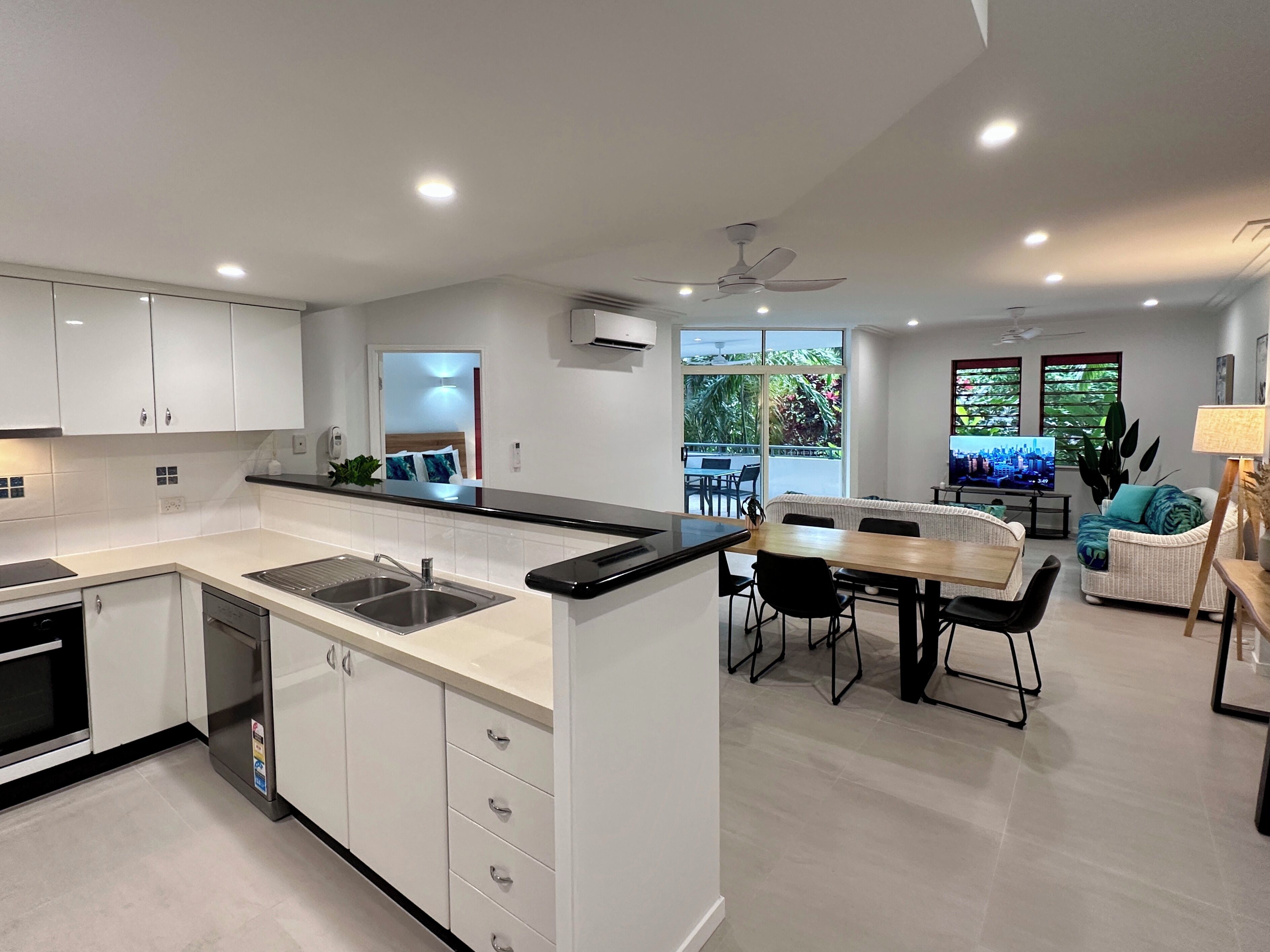 A modern kitchen with white cabinets, a black countertop, and a built-in sink. There is a dining area with a wooden table and chairs, and a sofa with a view of the outdoors.