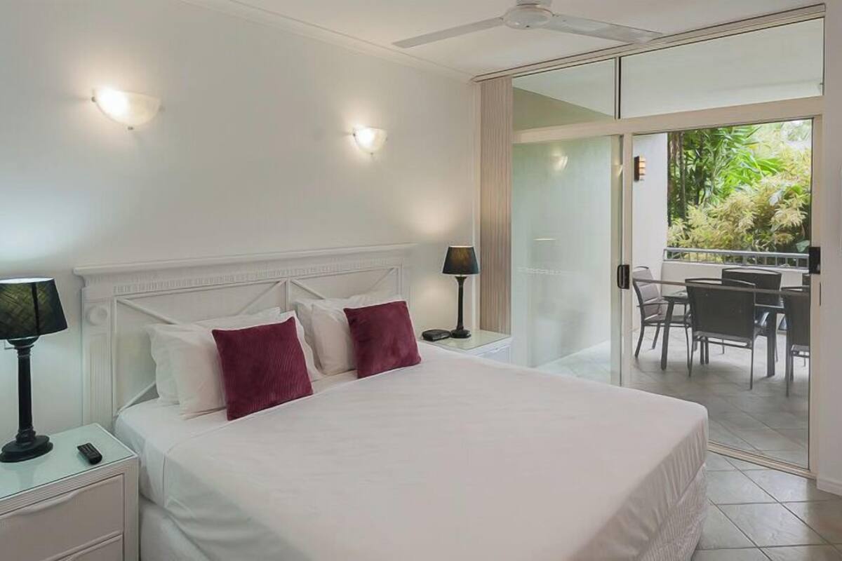 A hotel room with a large bed, two bedside lamps, a flat-screen TV, and a balcony with a table and chairs.