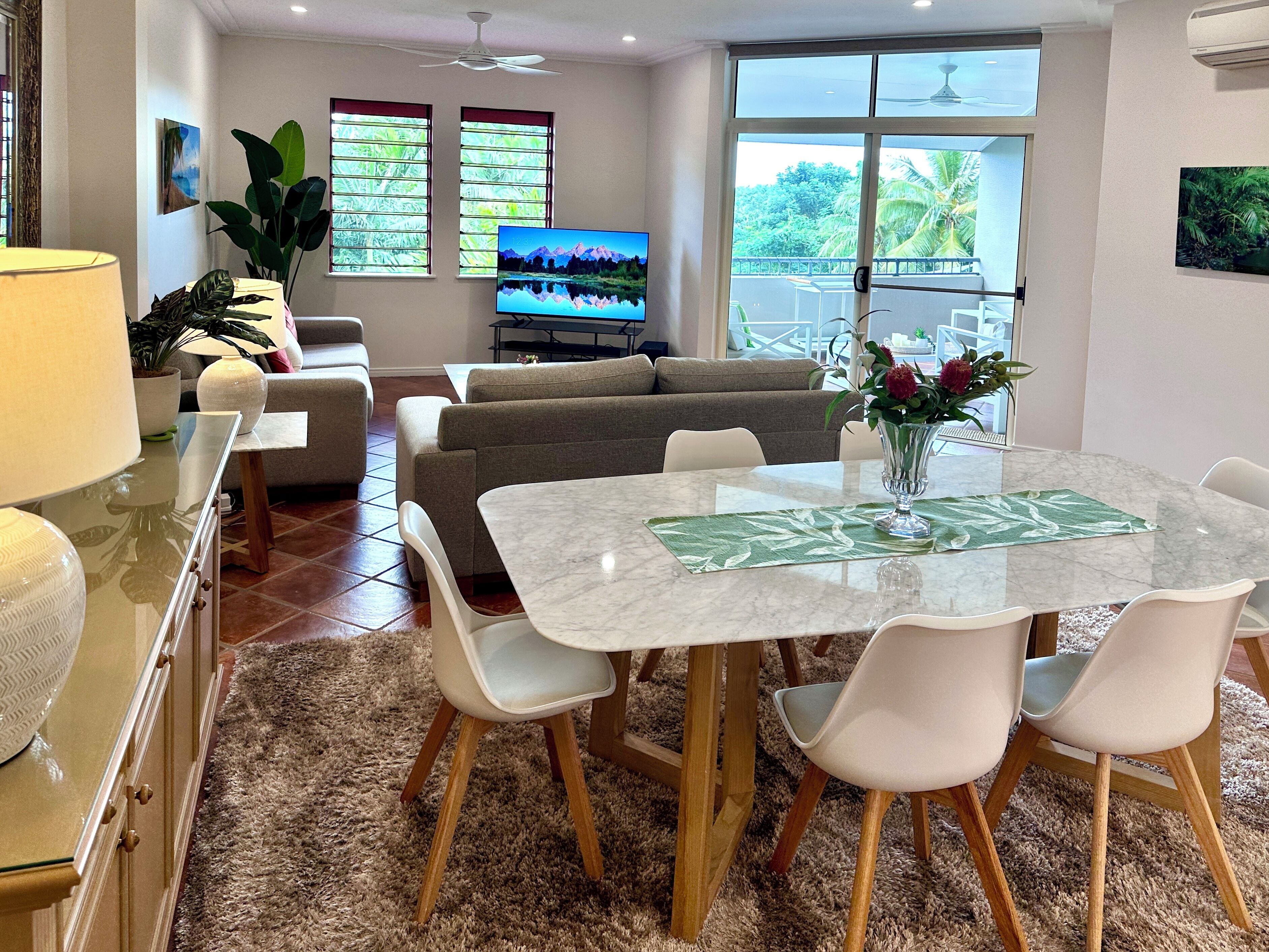 A modern living room with a dining area, a sofa set, a TV, and a large window with a view of greenery.