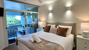 A hotel room with a large bed, a ceiling fan, a balcony with a view of greenery, and a small desk with a lamp. - Oasis at Palm Cove (Palm Cove)