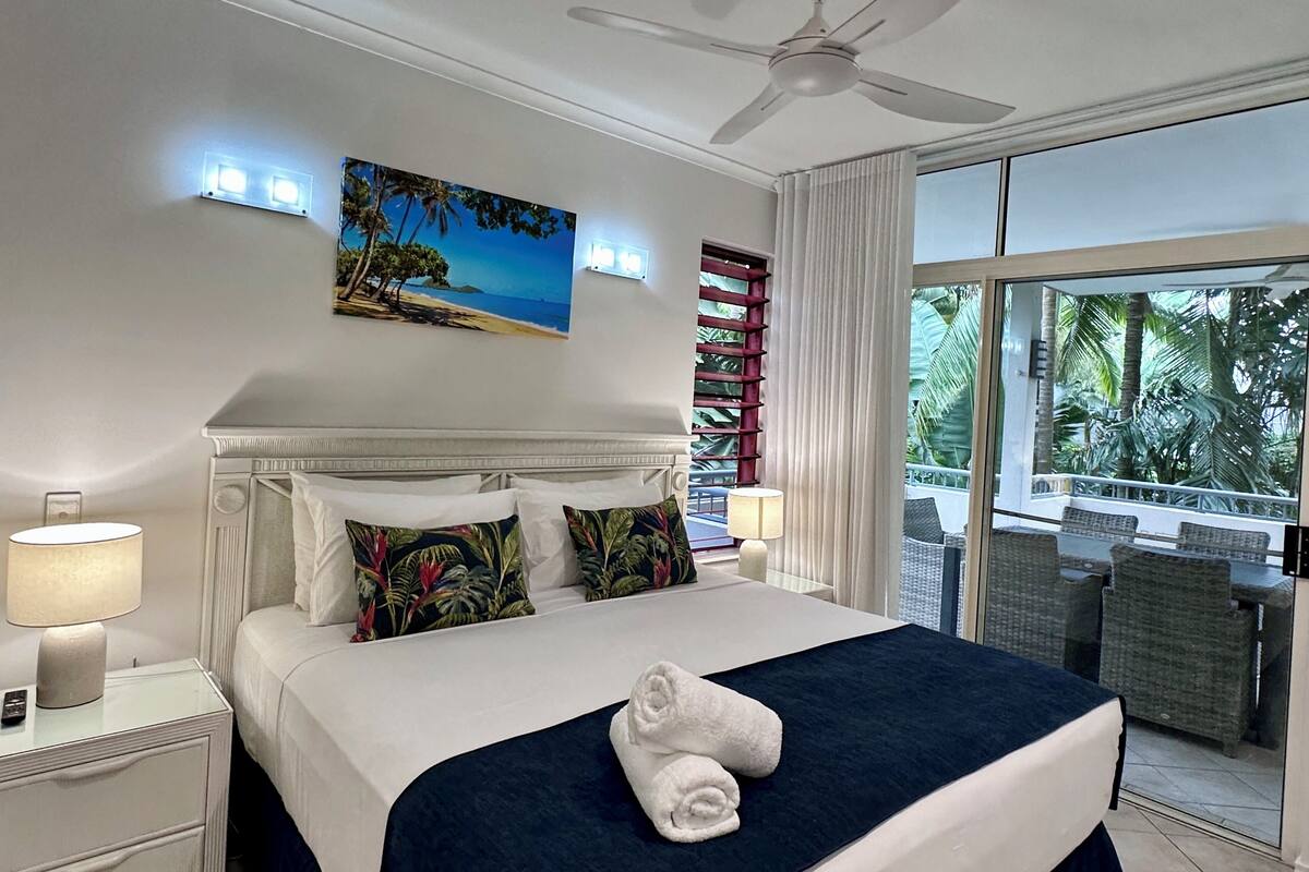 A hotel room with a bed, two bedside lamps, a ceiling fan, a large window with a view of palm trees, and a painting of a beach scene.