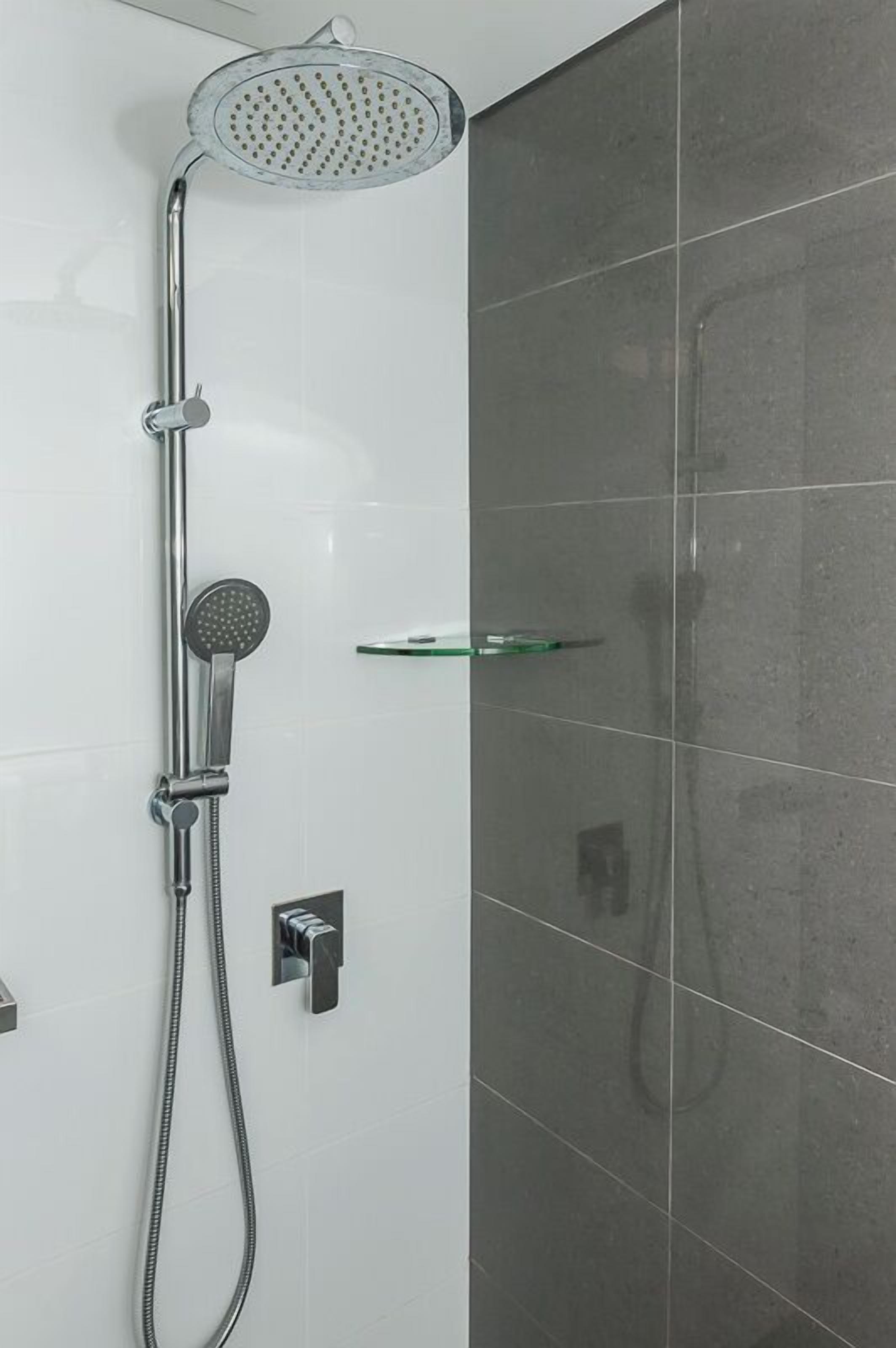 A modern shower with a handheld showerhead and a fixed showerhead, a glass shelf, and a control panel.