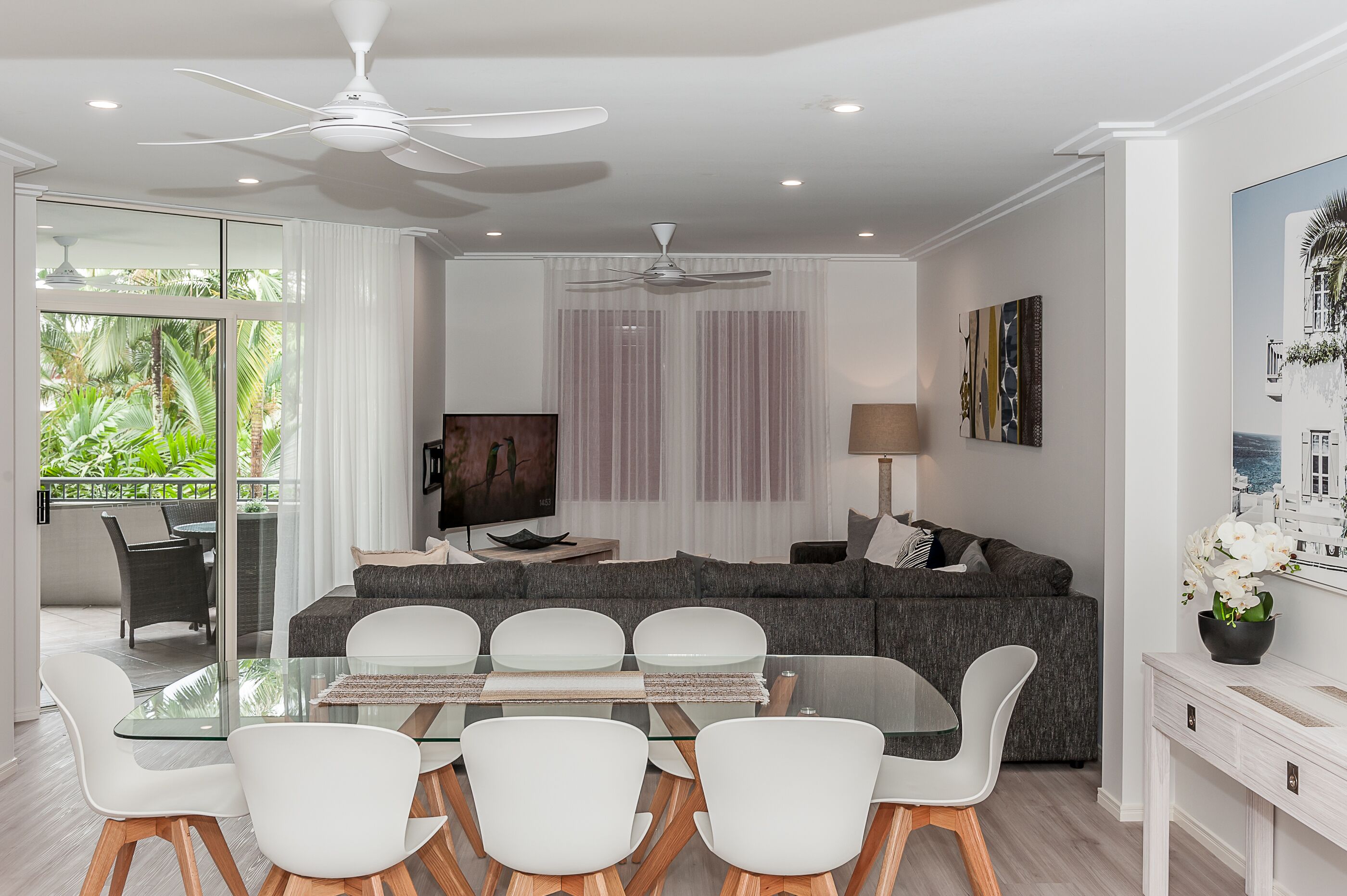 A modern living room with a glass dining table, white chairs, a grey sofa, and a ceiling fan.