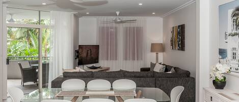 A modern living room with a glass dining table, white chairs, a grey sofa, and a ceiling fan.