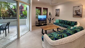 A living room with a TV, sofa, coffee table, and a balcony with a view of greenery. - Oasis at Palm Cove (Palm Cove)