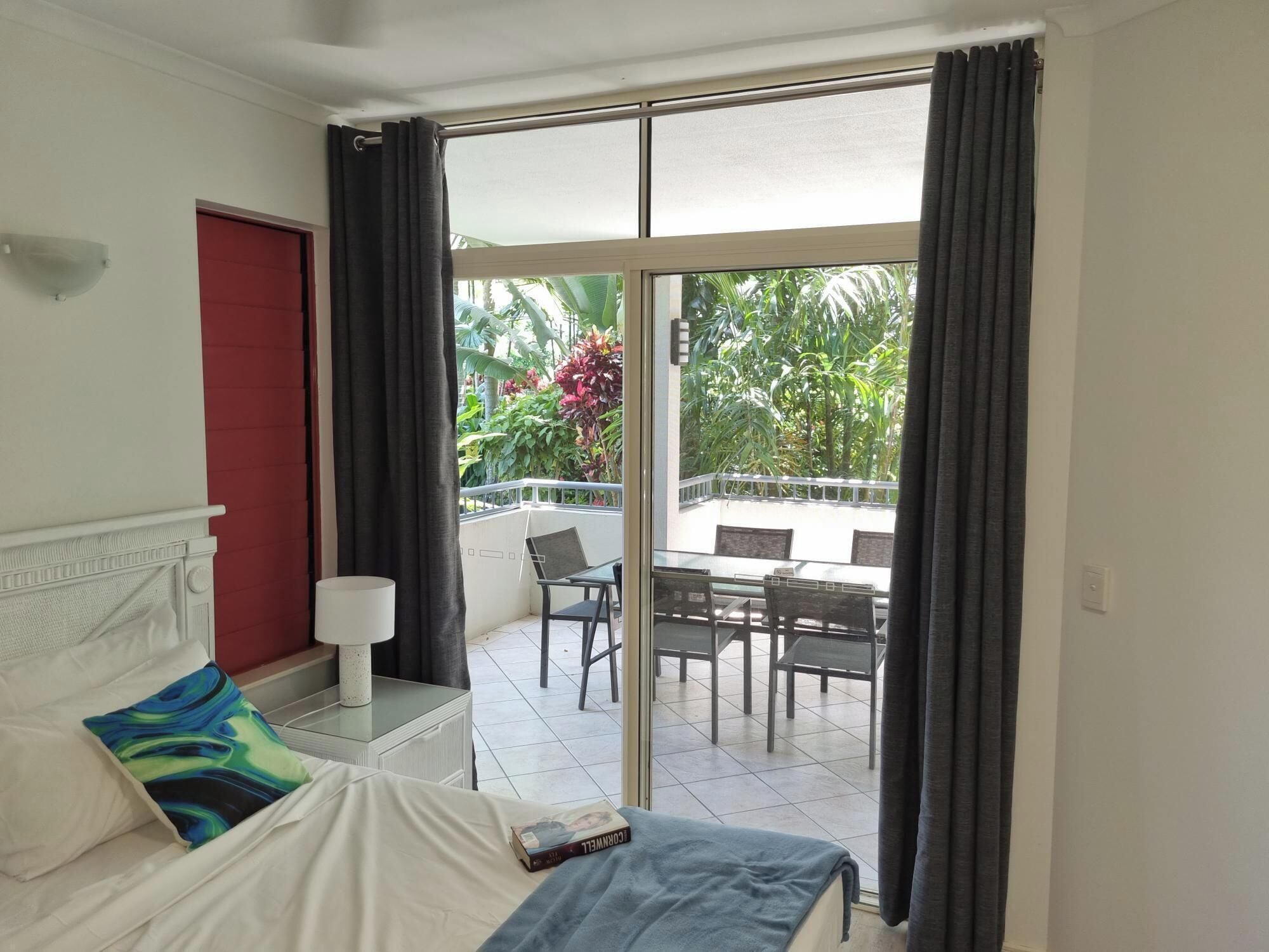 A hotel room with a bed, a nightstand, a lamp, a window with curtains, and a view of an outdoor balcony with chairs and plants.