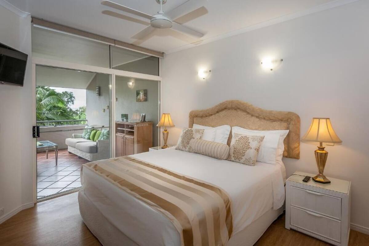 A bedroom with a bed, two bedside lamps, a ceiling fan, and a view of a balcony.