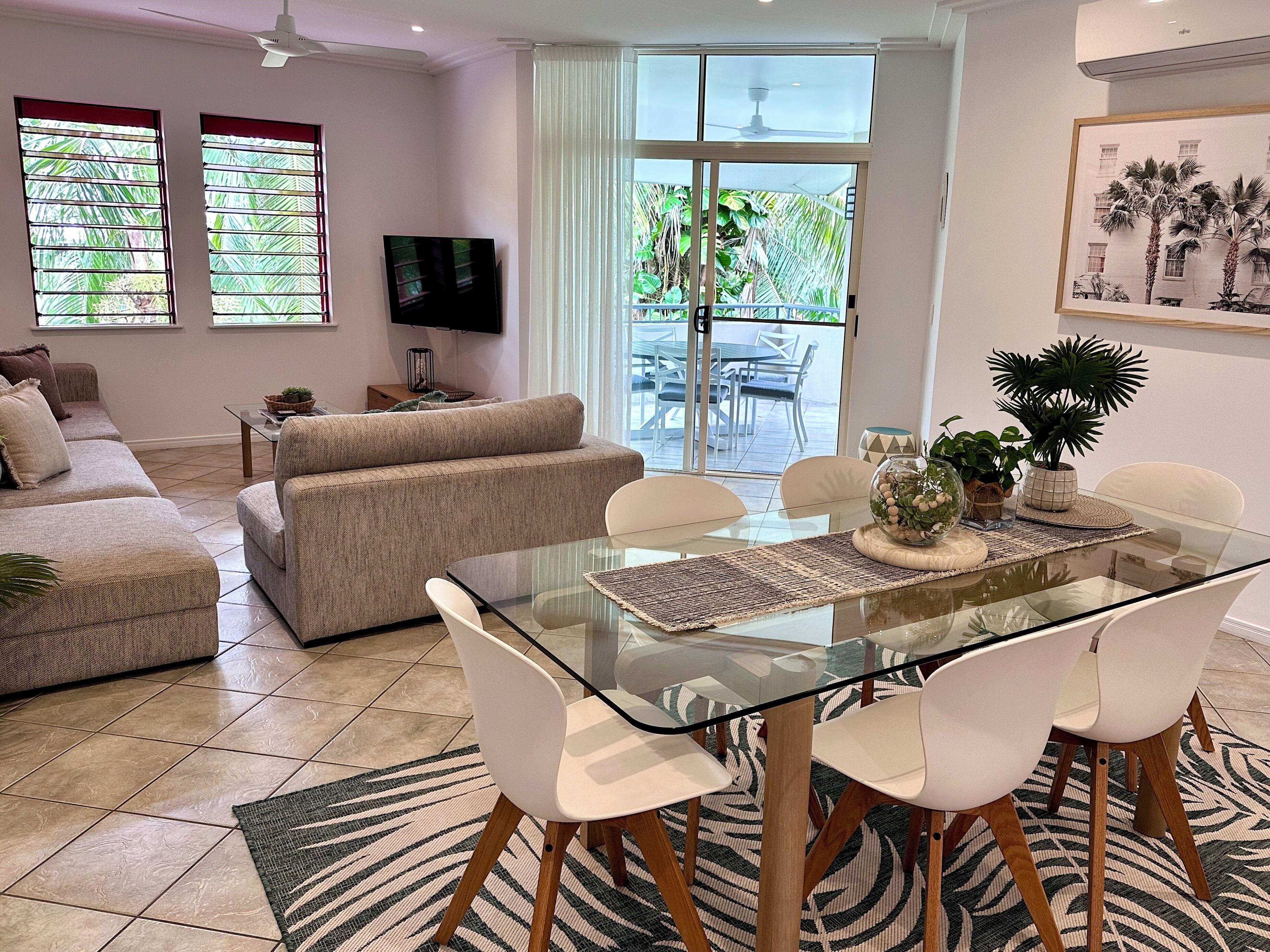 Una sala de estar moderna con una mesa de comedor de vidrio, sillas blancas, un sofá beige y un televisor en la pared.