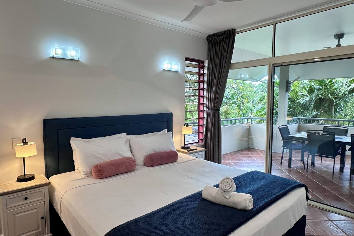 A hotel room with a bed, nightstand, lamp, and a view of the outdoors.