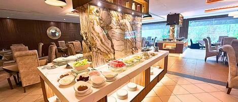 Disposição de buffet com diversos pratos, uma parede de detalhes em mármore e uma área de estar com cadeiras e mesas.