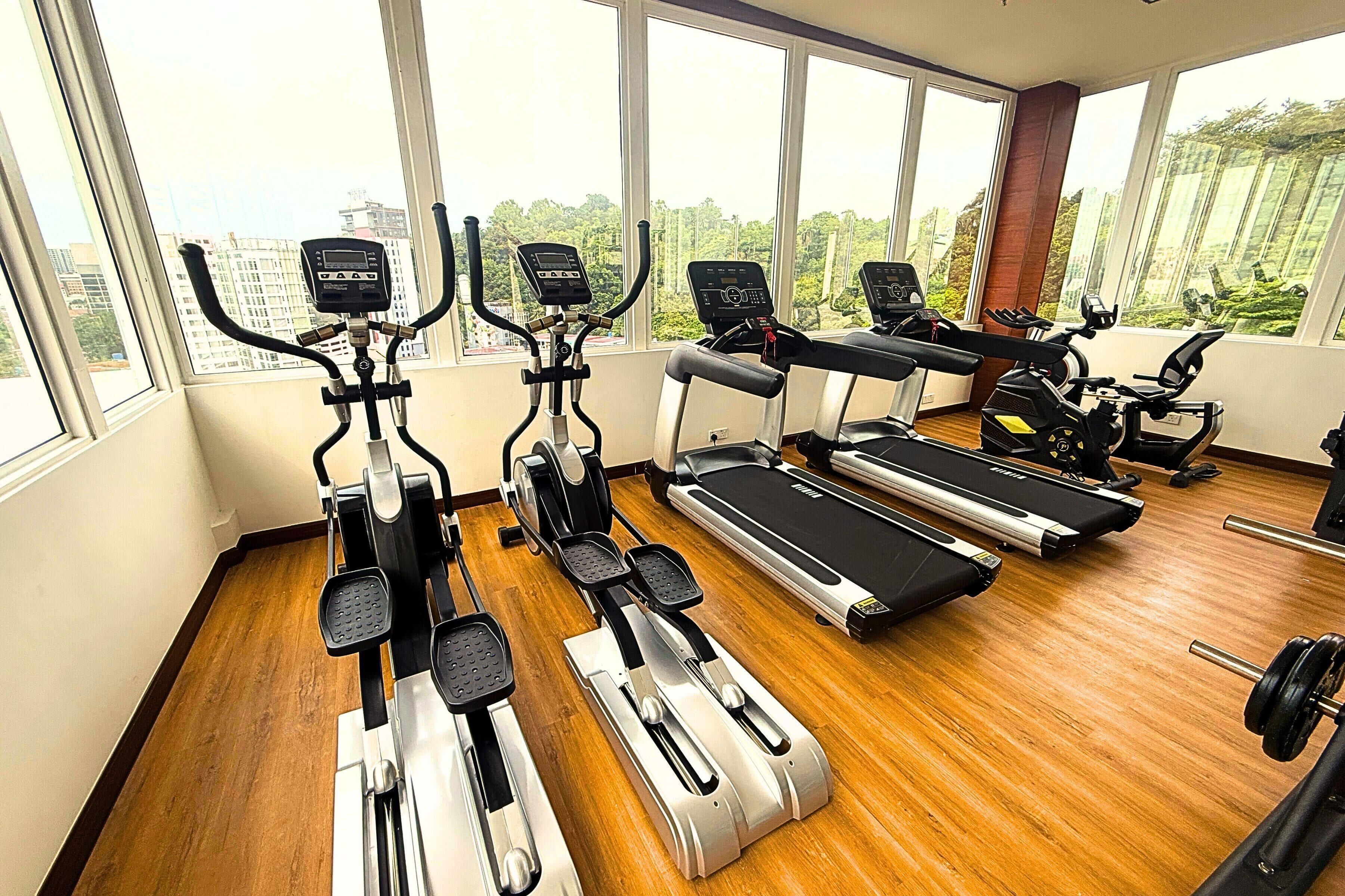 A gym with cardio machines and a treadmill, large windows, and a wooden floor.
