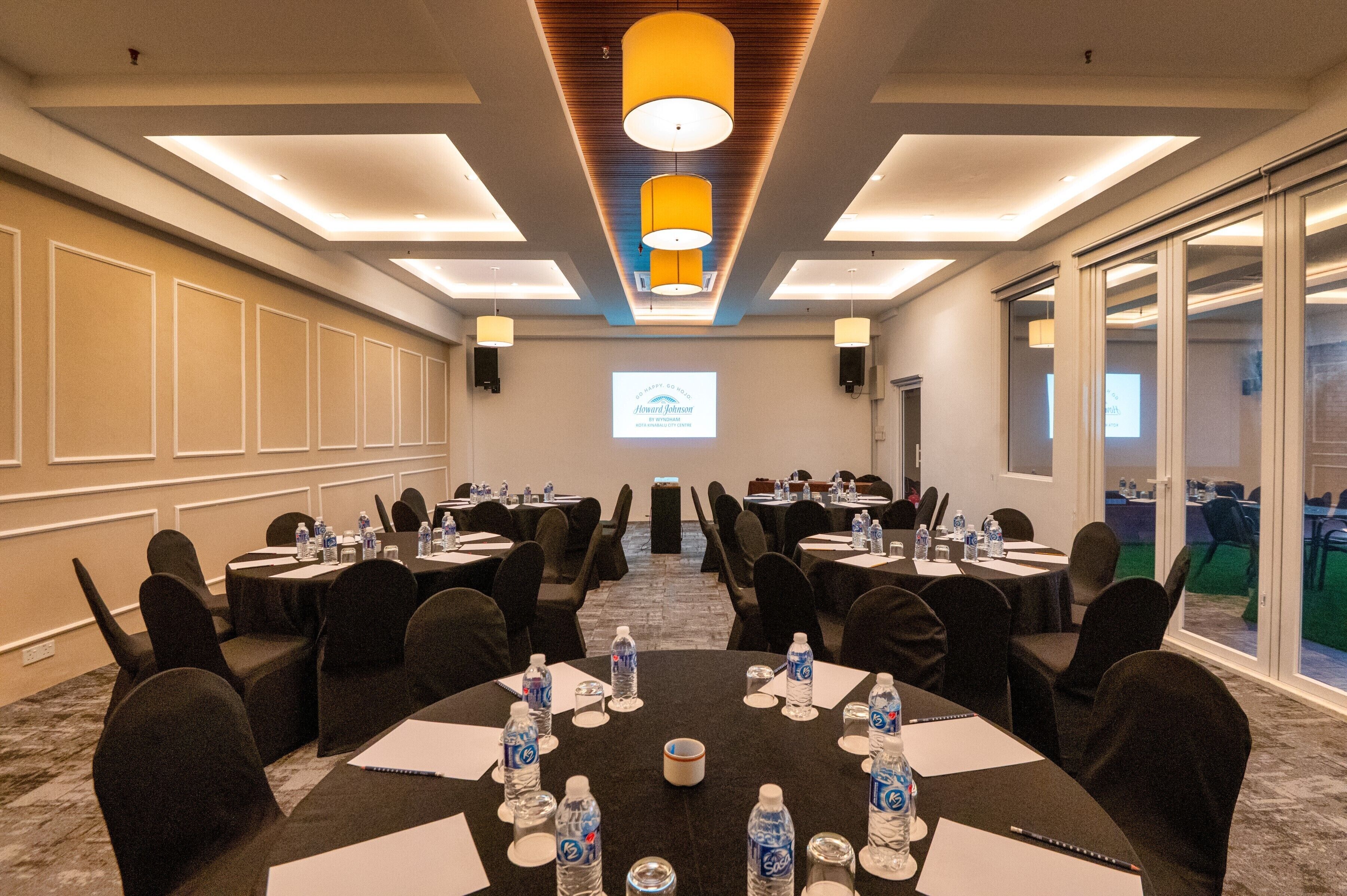A conference room with round tables, black chairs, water bottles, and a projector screen.