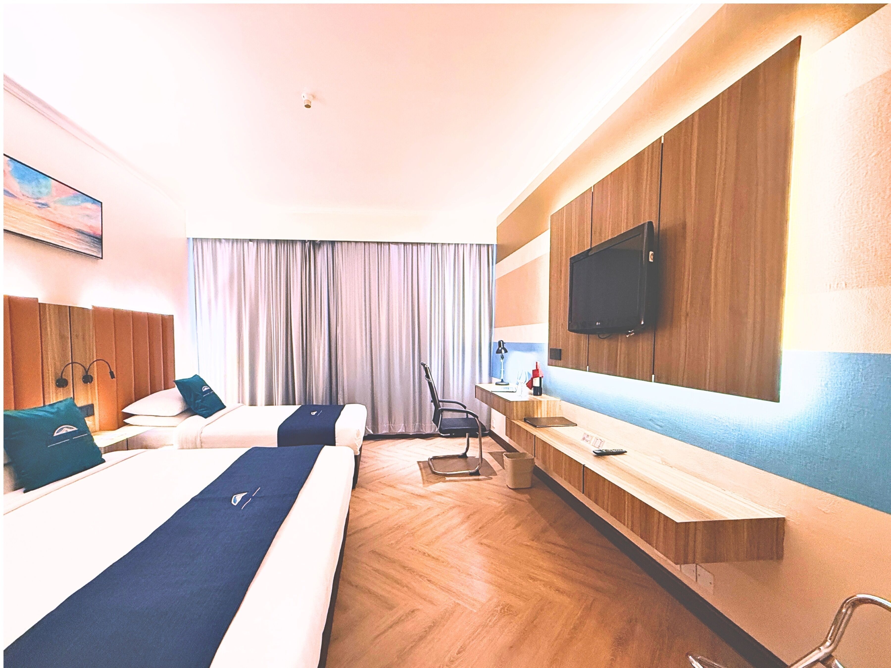 Habitación de hotel con dos camas, un escritorio, una silla, un televisor y un ventanal con cortinas.