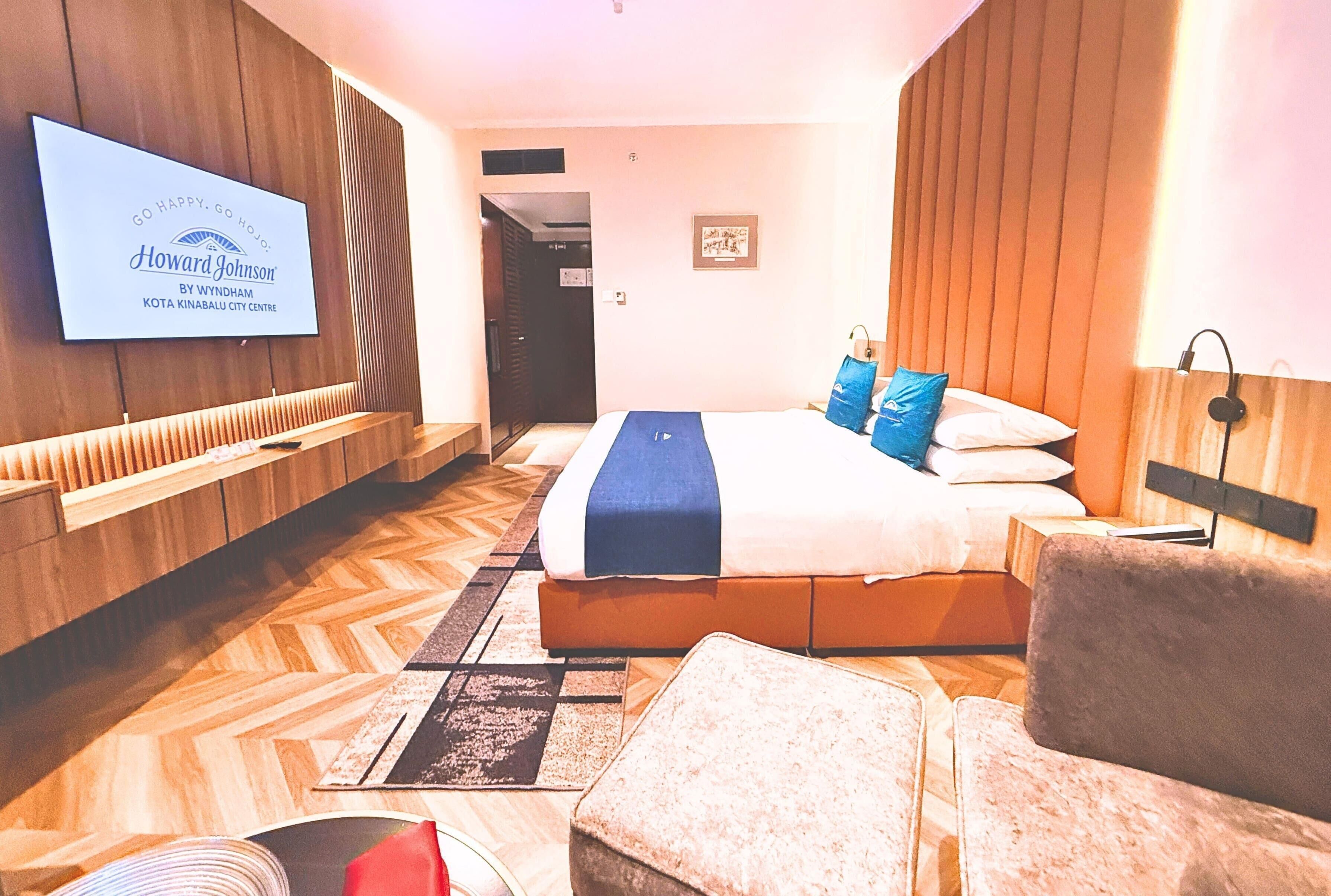 A hotel room with a bed, a TV mounted on the wall, a wooden headboard, and a seating area with a chair and ottoman.