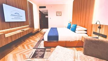 A hotel room with a bed, a TV mounted on the wall, a wooden headboard, and a seating area with a chair and ottoman.