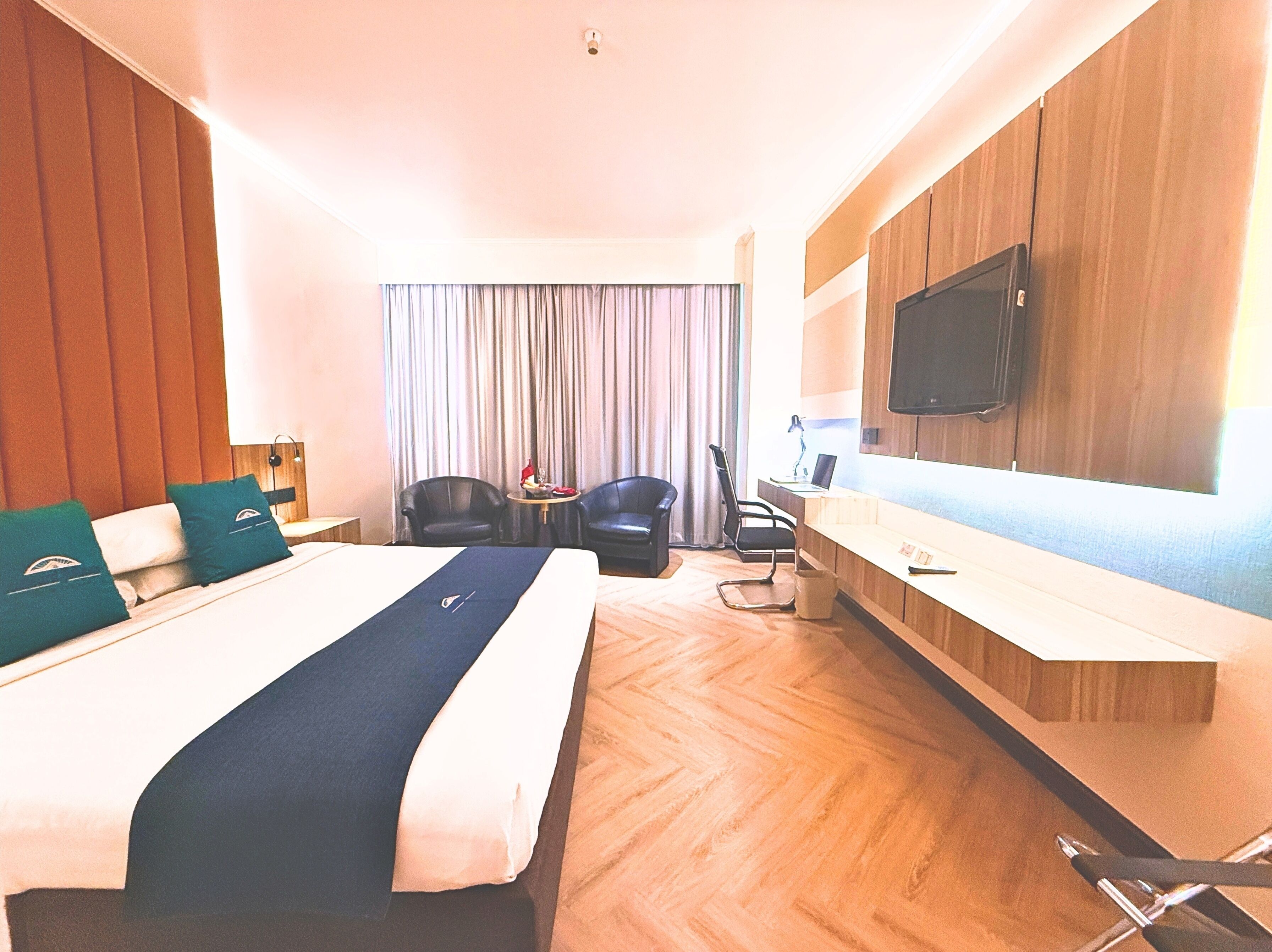 A hotel room with a large bed, a desk, two chairs, a TV, and a window with curtains.