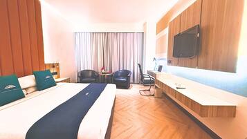 A hotel room with a large bed, a desk, two chairs, a TV, and a window with curtains.