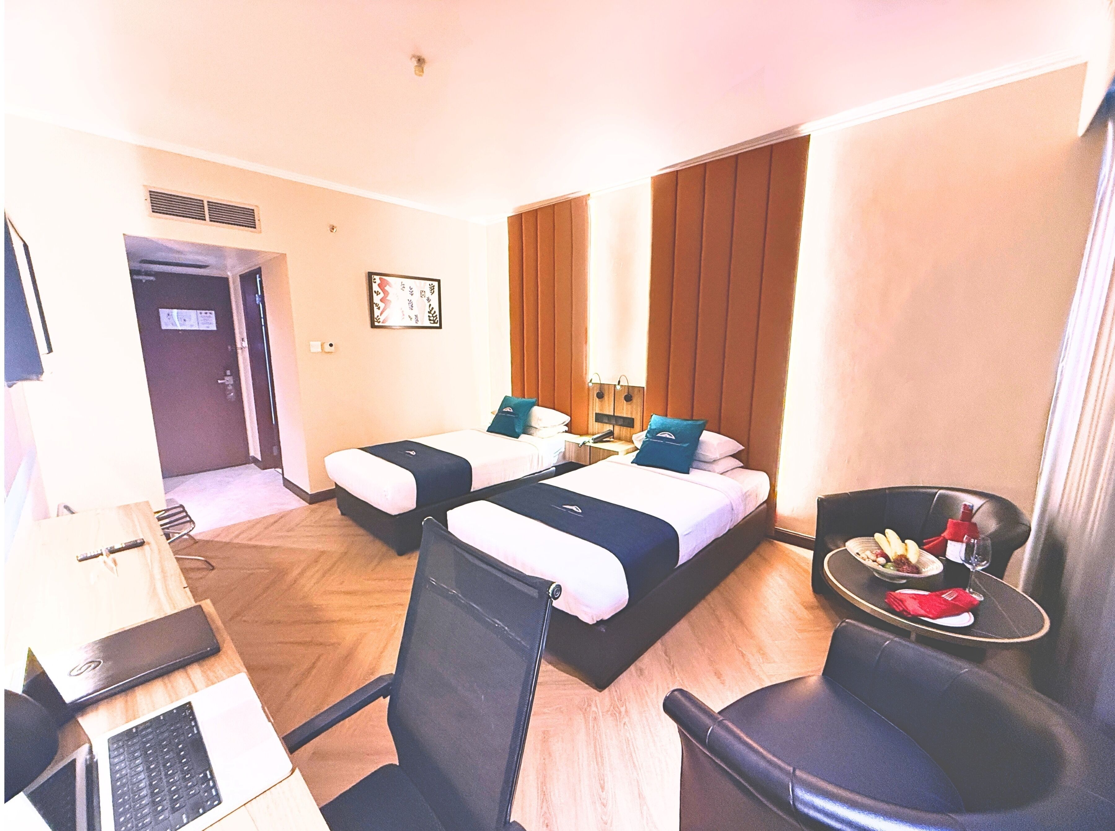 A hotel room with two beds, a desk with a laptop, a chair, and a small table with food and drink.