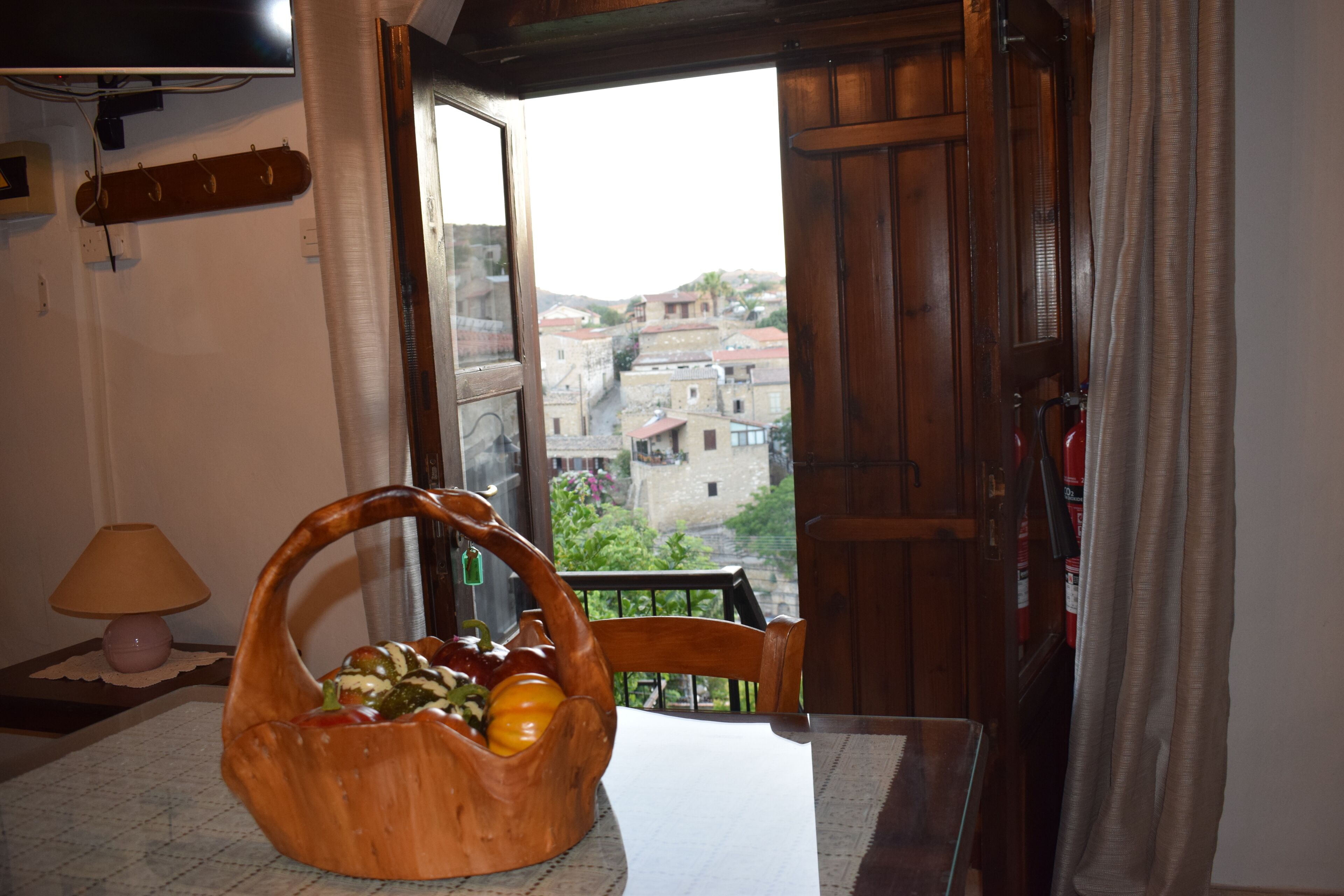 A room with a wooden table, a wicker fruit basket, a lamp, and a view of buildings through an open door.
