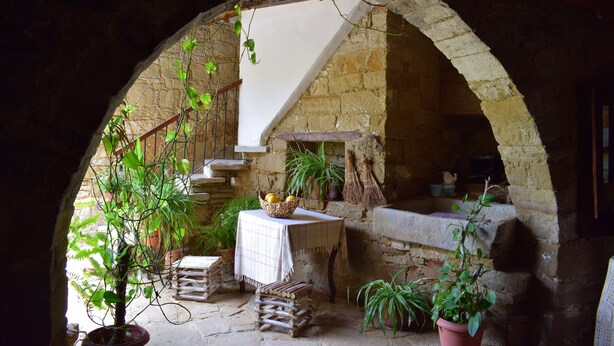 Un'antica arcata in pietra introduce un ambiente accogliente con un tavolo, piante in vaso e un camino in pietra.