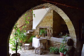 A rustic stone archway leading to a cozy interior with a table, potted plants, and a stone fireplace. - Vasilopoulos House (Tochni)