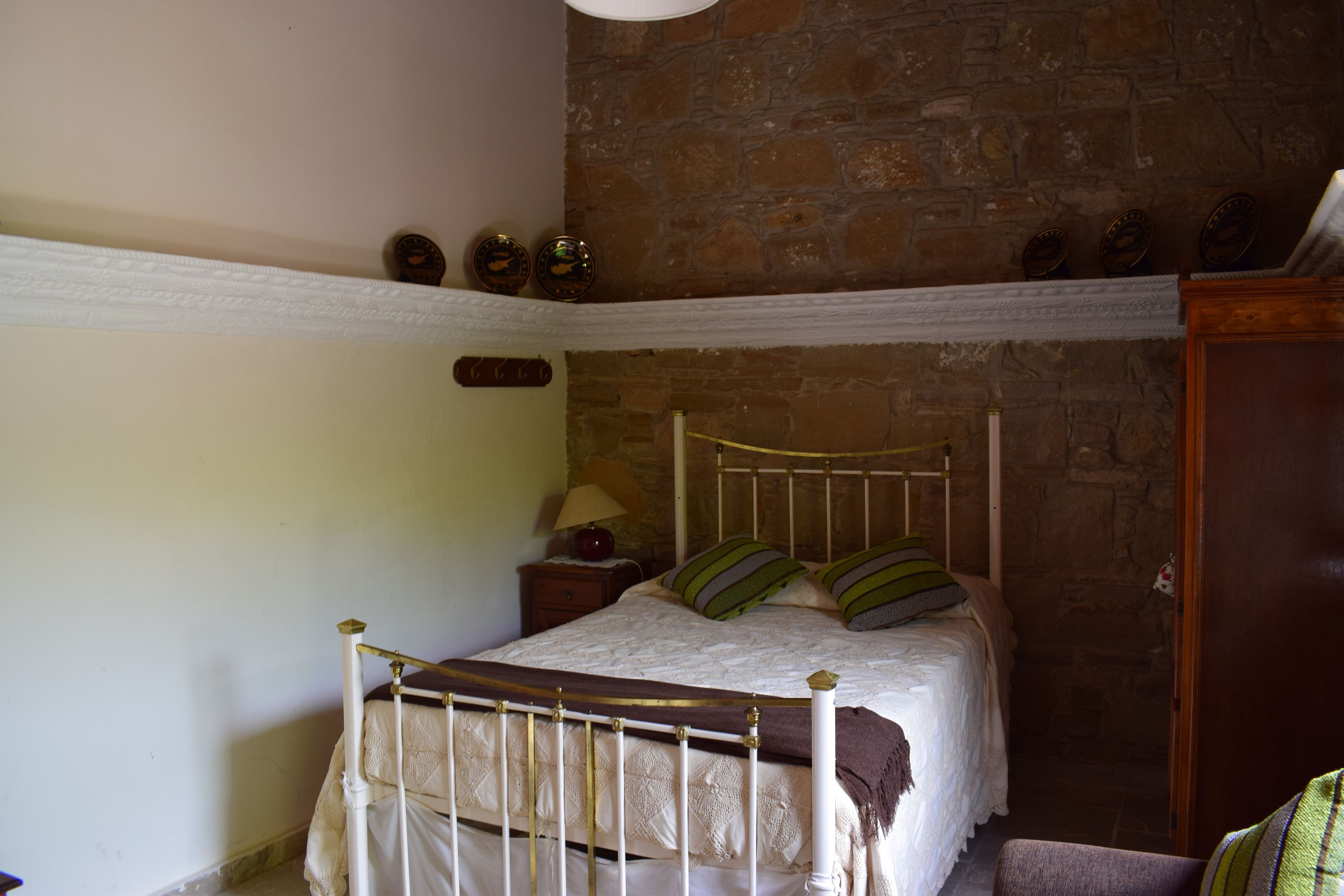 A bedroom with a stone wall, a wooden bed frame, a nightstand with a lamp, and a shelf with decorative items.
