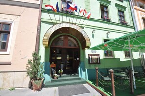 A building with a green facade, a sign reading "BARBARA," and multiple flags. There are outdoor tables and chairs, and a potted plant near the entrance. - GARNI - Barbakan Hotel (Levoca)