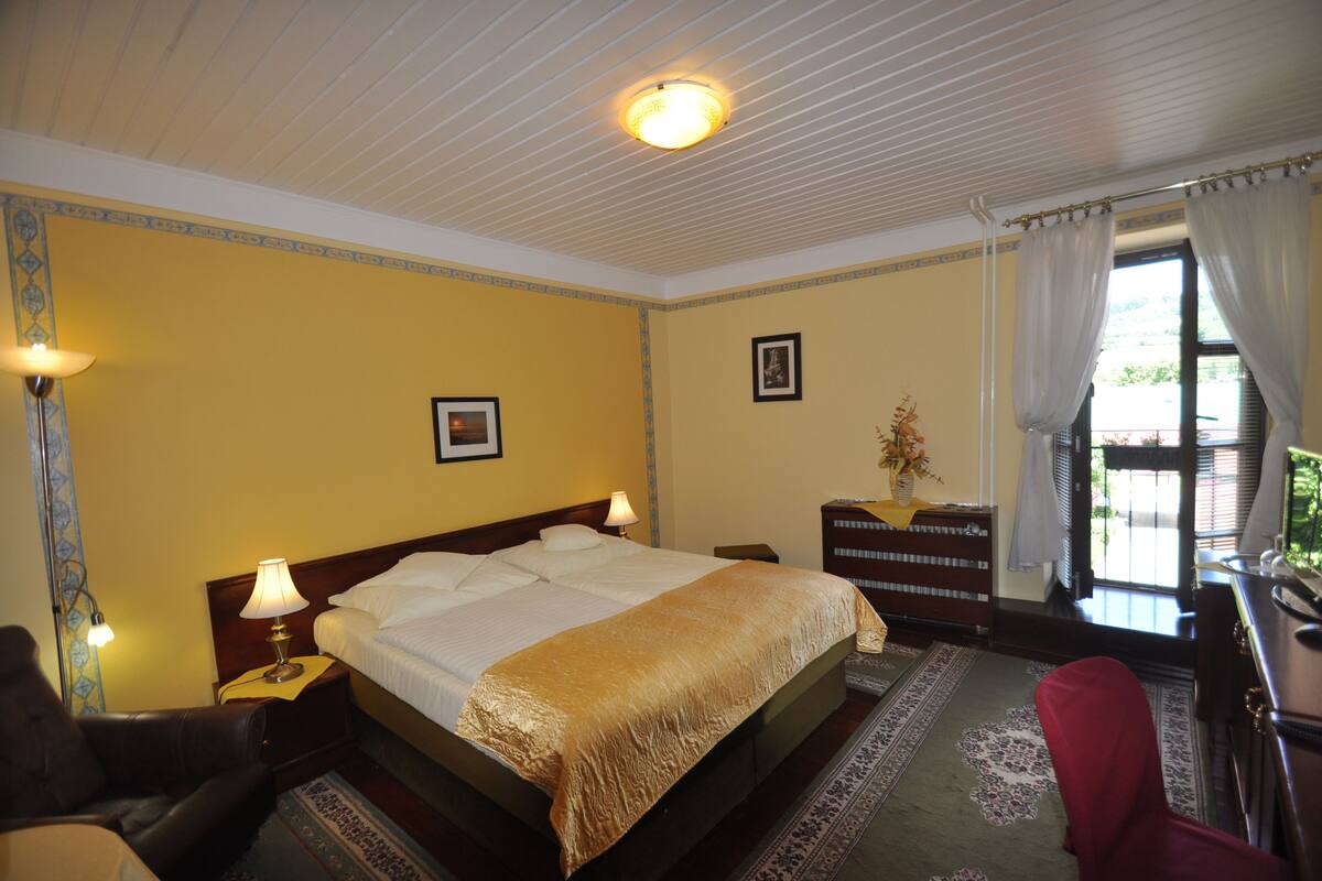 Een hotelkamer met een groot bed, twee bedlampen, een houten dressoir, een rode stoel en uitzicht op het groen door een raam met gordijnen.