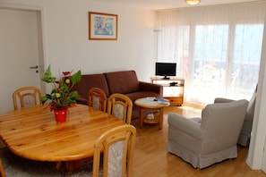 A living room with a wooden dining table, a brown sofa, a television on a stand, and a large window with sheer curtains. - Résidence Chamossaire (Leysin)