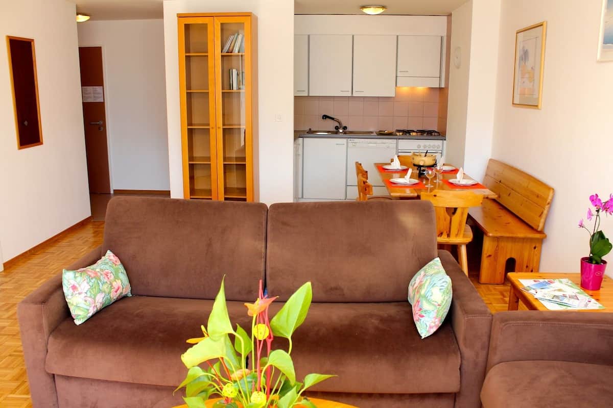 Ein Wohnzimmer mit braunem Sofa, einem Esstisch mit Stühlen, einer Küchenzeile und einem Bücherregal.