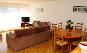 A living room with a brown sofa, wooden dining table, and chairs. A television is on a stand, and there are potted plants on the table. - Résidence Chamossaire (Leysin)