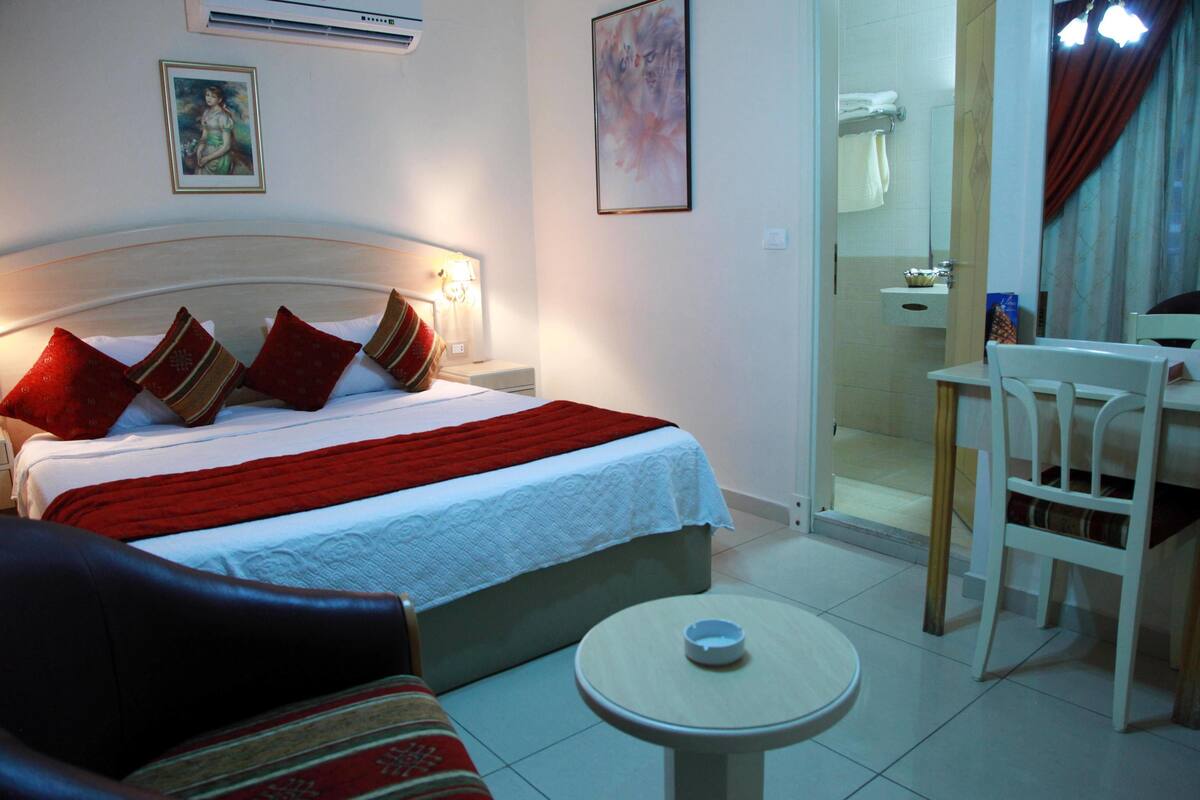 Habitación de hotel con cama, una mesita, una silla, un sofá y un baño visible a través de una puerta abierta.
