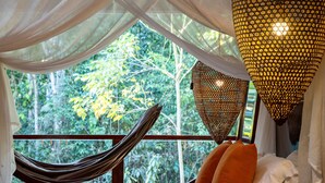 Un lit avec un hamac, une suspension lumineuse et une vue sur les arbres par la fenêtre.