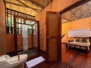 A wooden-framed room with a bed, a toilet, and a door leading to an outdoor area.
