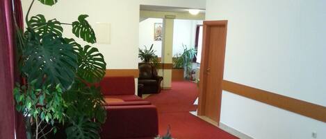 Un vestíbulo de hotel con alfombra roja, un mostrador de recepción de madera, una zona de estar con sofás rojos y plantas verdes grandes.