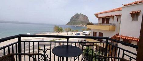 A balcony with wicker chairs and a small table, overlooking a beach and a rocky island.