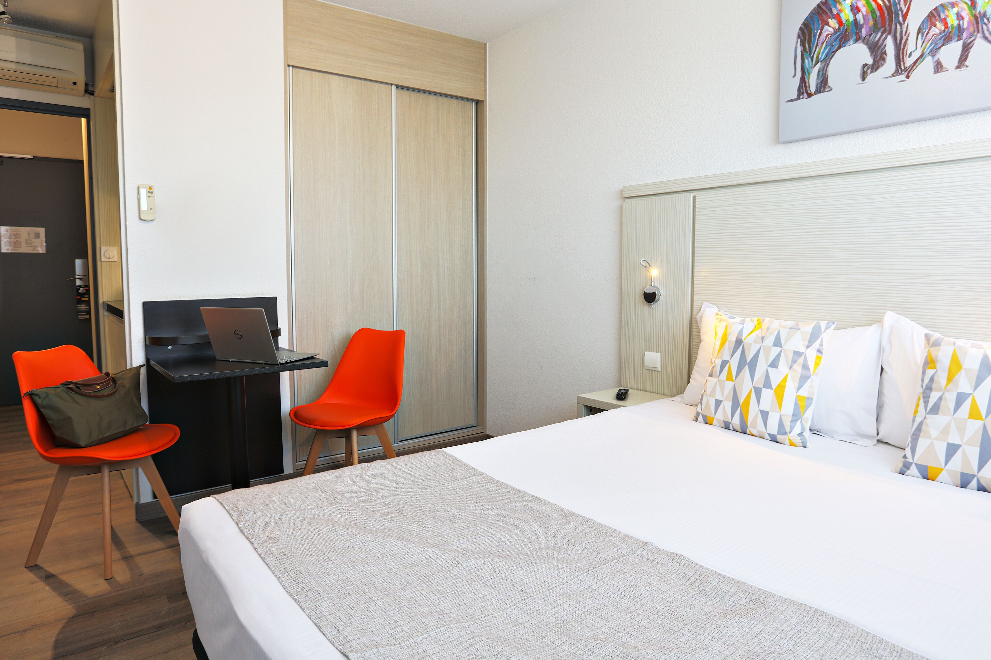 Una habitación de hotel con una cama, un escritorio con una computadora portátil, dos sillones naranjas y un cuadro en la pared.